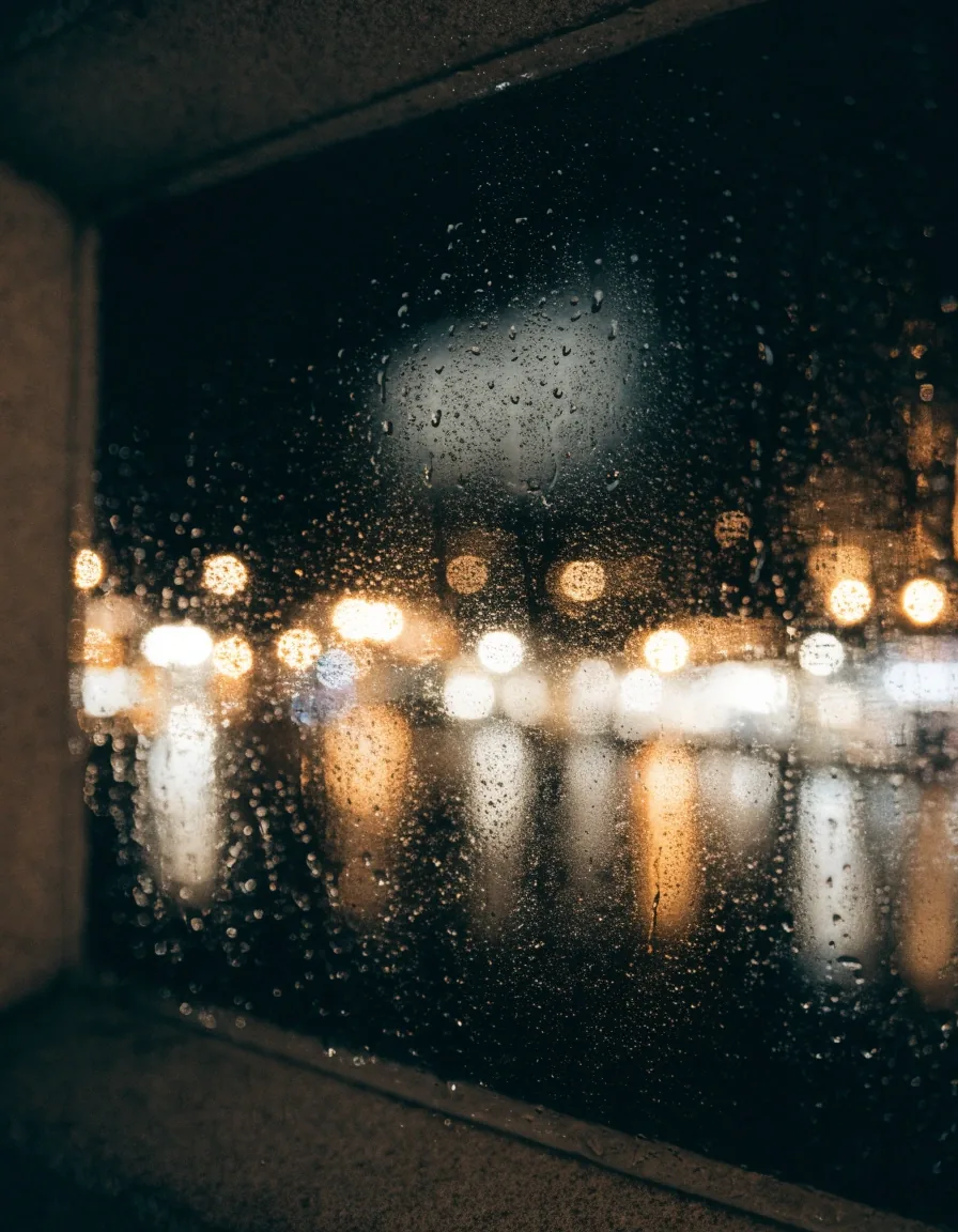 Superficie de vidrio con lluvia y luces urbanas difusas al fondo, sin figuras ni rótulos